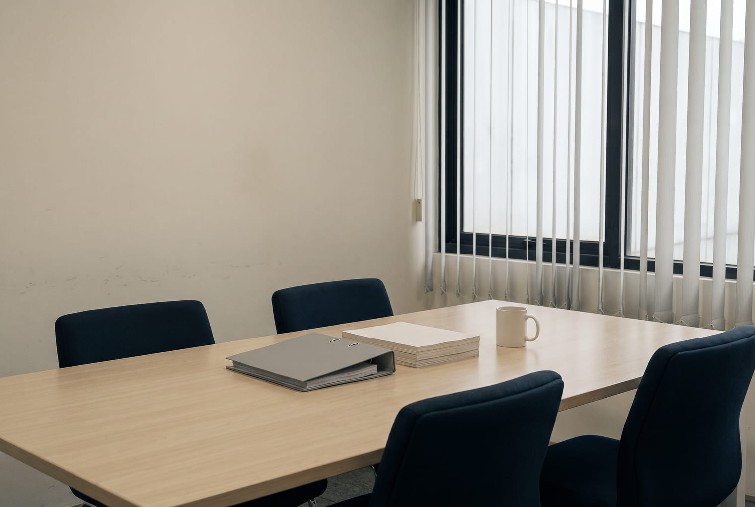 Ruang rapat kantor Artha Consulting: meja kayu terang dengan empat kursi, satu ring binder tertutup dan setumpuk kertas, di depan jendela berkerai vertikal.